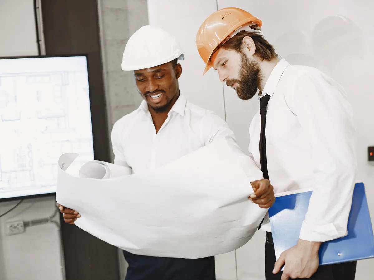 Two engineers in hard hats unrolling a drawing in an office, with a floor plan on a screen behind them.