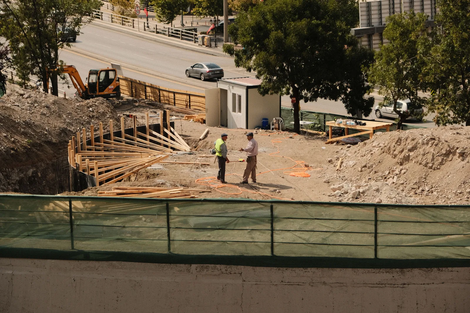 Two workers, one in a hi-vis vest, talking on a roadside excavation with timber formwork and an excavator behind them.